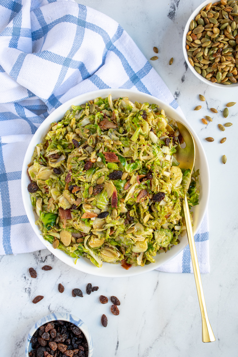 White serving bowl and gold serving spoon with a warm brussel sprout bacon salad garnished with raisins and pumpkin seeds.