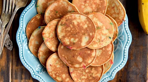 Mini Whole Wheat Banana Pancakes on a blue plate surrounded by sprinkles, a cup of flour, and a banana