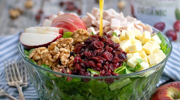 Apple Cider Vinaigrette being poured onto an Apple Cranberry Salad with turkey, walnuts, and cheddar cheese