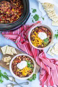 Two bowls and a crockpot full of Three Bean Vegetarian Chili on a table top with garnishes, saltine crackers, a kitchen towels.