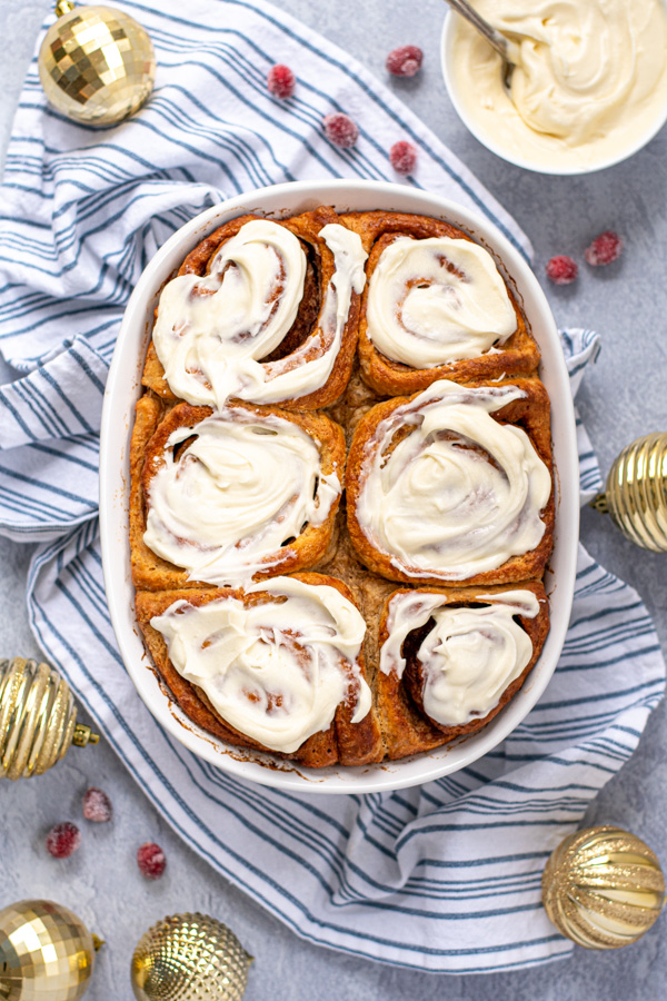 Freshly made cinnamon rolls covered in cream cheese frosting sitting next to golden Christmas ornaments and sugared cranberries.
