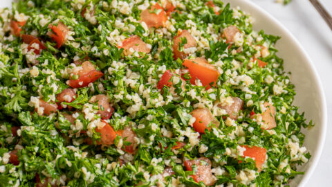 A large white bowl filled with tabbouleh salad with a spoonful out on the side with parsley leaves falling off.