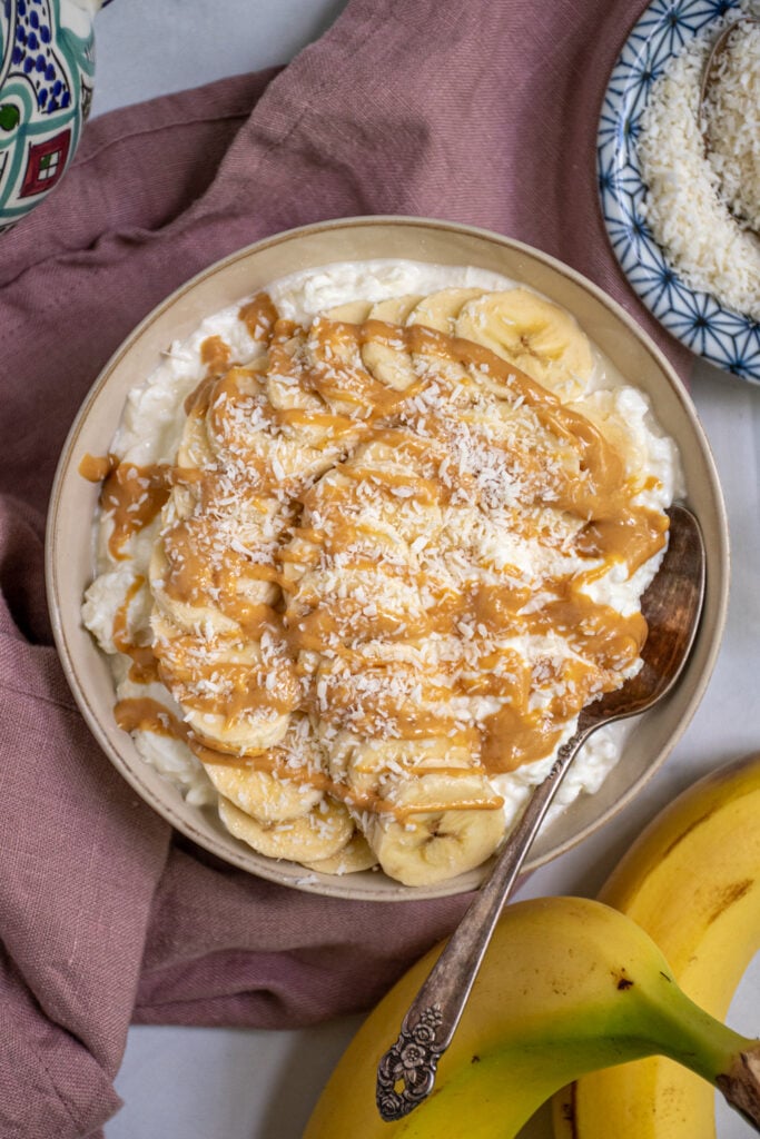 Close up of a bowl filled with cottage cheese and topped with banana slices and drizzled peanut butter with shredded coconut flakes.