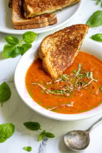 A white bowl filled with roasted red pepper and tomato soup garnished with fresh basil and a toasted grilled cheese being dunked into the soup.