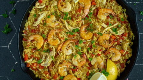 Overhead view of a black pan full of chicken and shrimp paella showing off yellow rice, chicken, red bell pepper, and cooked shrimp garnished with fresh parsley and lemon wedges.