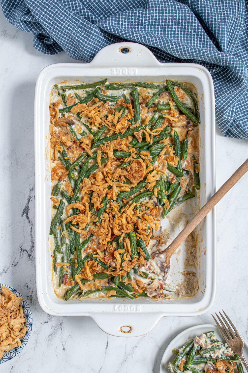 Overhead view of a white casserole dish with homemade green bean casserole with a serving spoon and some on a plate next to it.