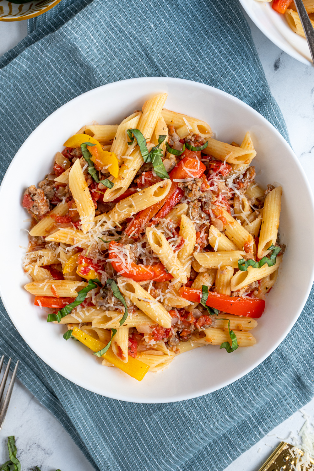 An overhead view of a white bowl filled with penne pasta, sausage, and bell peppers and garnished with parmesan cheese.