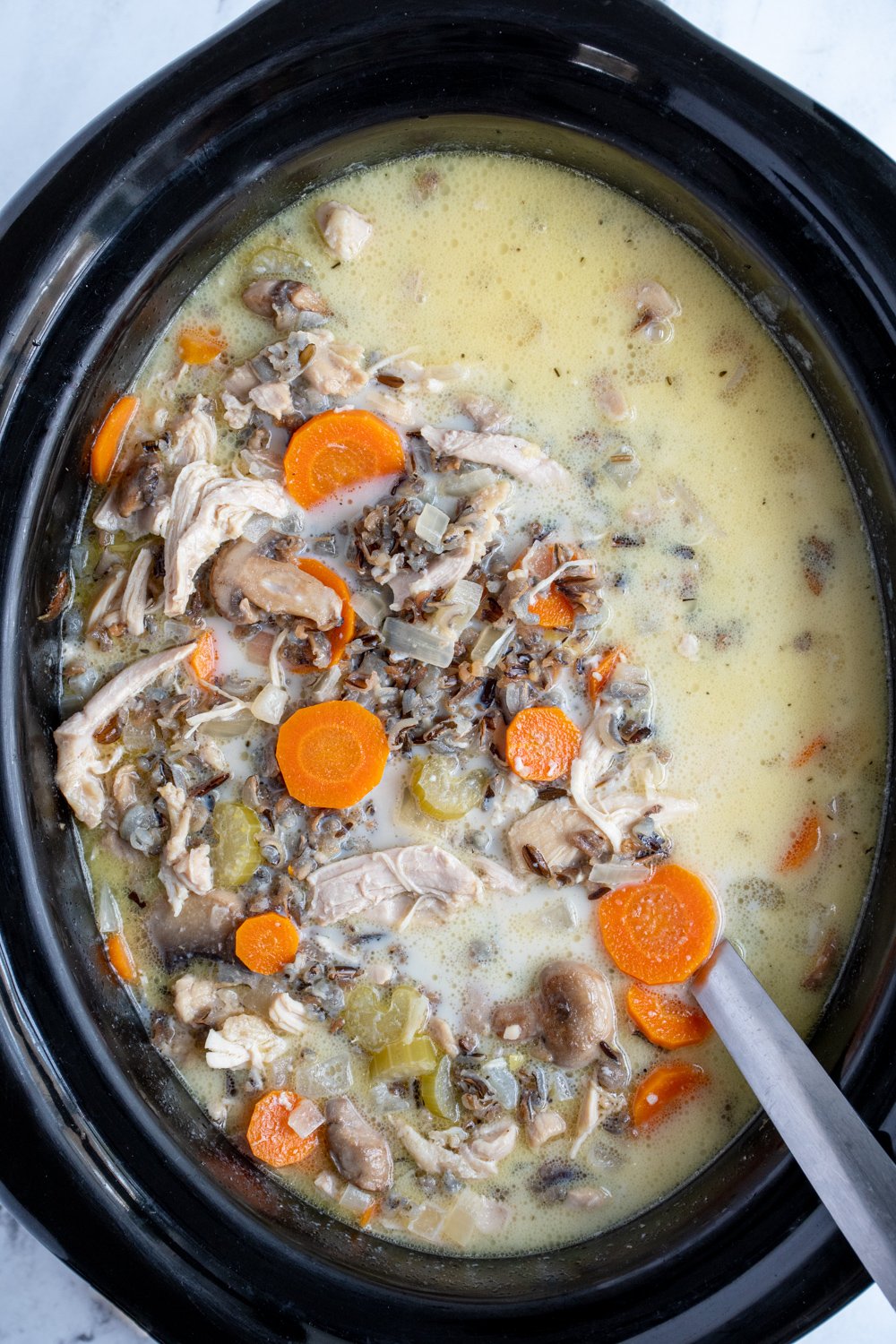 Black crockpot filled with chicken wild rice soup with a ladle showing off wild rice, carrots, celery, and chicken.