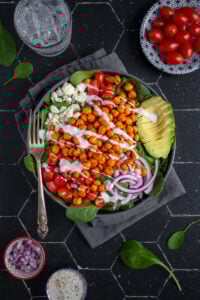 Overhead view of a black bowl on a black background, the bow is filled with buffalo chickpea salad, garnished with a ranch drizzle, avocado, red onion, and tomatoes.