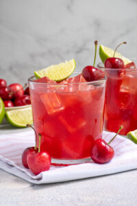 A glass filled with ice and bright red cherry margarita topped with a fresh cherry and lime wedge.