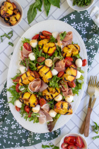 Overhead view of a large white platter filled with mixed greens, grilled peaches, tomatoes, mozzarella, and prosciutto making a Grilled Peach and Mozzarella Salad.