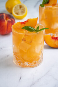 A glass full of Bourbon Peach Smash an orange colored cocktail garnished with a peach slice and basil leaves.