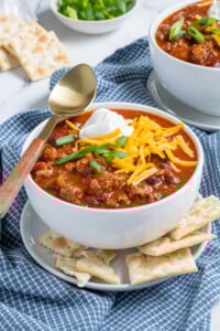 Bowl of classic chili with ground beef and beans topped with sour cream, cheese, and green onion with saltine crackers.