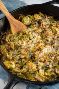 A skillet filled with cheesy brussel sprouts au gratin with a wood spoon scooping some up.