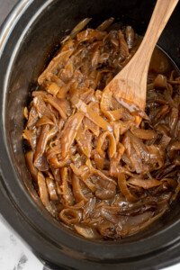 A black crockpot with slow cooker caramelized onions that a golden brown with a wooden spoon.