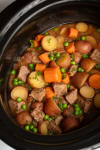 Crockpot full of beef stew filled with carrots, potatoes, chuck roast, and peas.