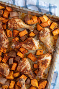A sheet pan with baked chicken legs and sweet potato chunks covered in seasoning.
