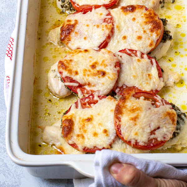 Hands holding a white baking dish containing pesto chicken bake, showing off melted mozzarella cheese on top.