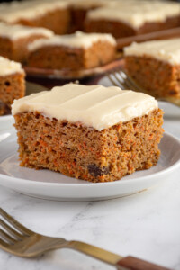 A white plate with a square cut piece of orange spiced easy carrot cake with orange flecks and raisins, topped with a cream cheese frosting.