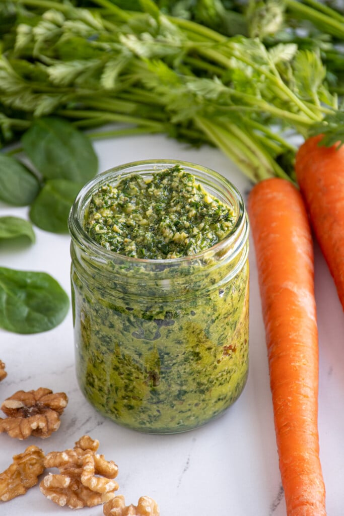 A glass jar filled with green carrot top pesto surrounded by a carrot with greens, spinach, and walnuts.