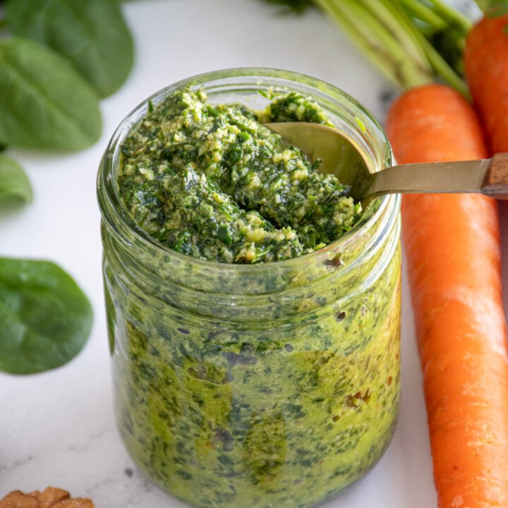A glass jar with a spoon scooping up green color carrot top pesto.