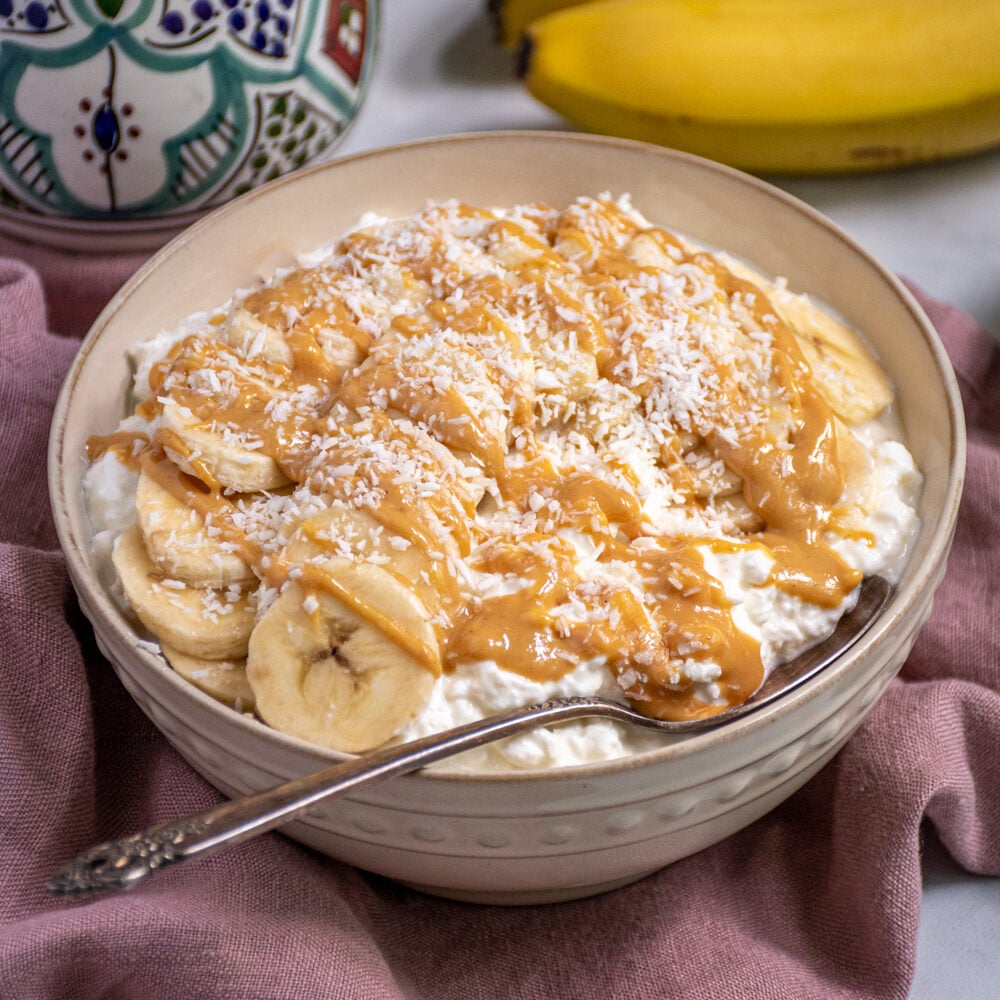White bowl gilled with cottage cheese and topped with banana slices, peanut butter, and shredded coconut.
