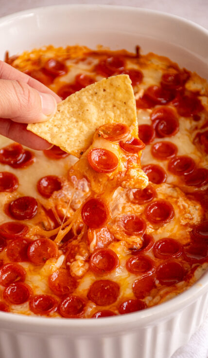A casserole dish filled with cottage cheese pizza dip covered in mini pepperoni with a tortilla chip being dipped and pulling along melted cheese.