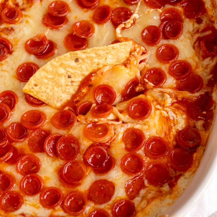 A white casserole dish with cheesy cottage cheese pizza dip covered in mini pepperoni and a tortilla chip scooped into the dip.