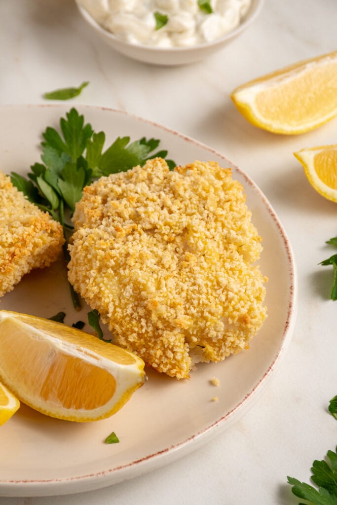 Plates with pieces of crispy baked breaded cod with lemons and fresh parsley.