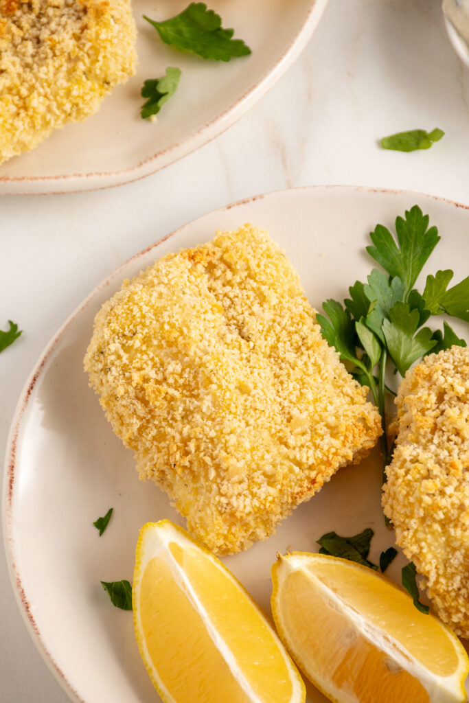 Plates with pieces of crispy baked breaded cod with lemons and fresh parsley.