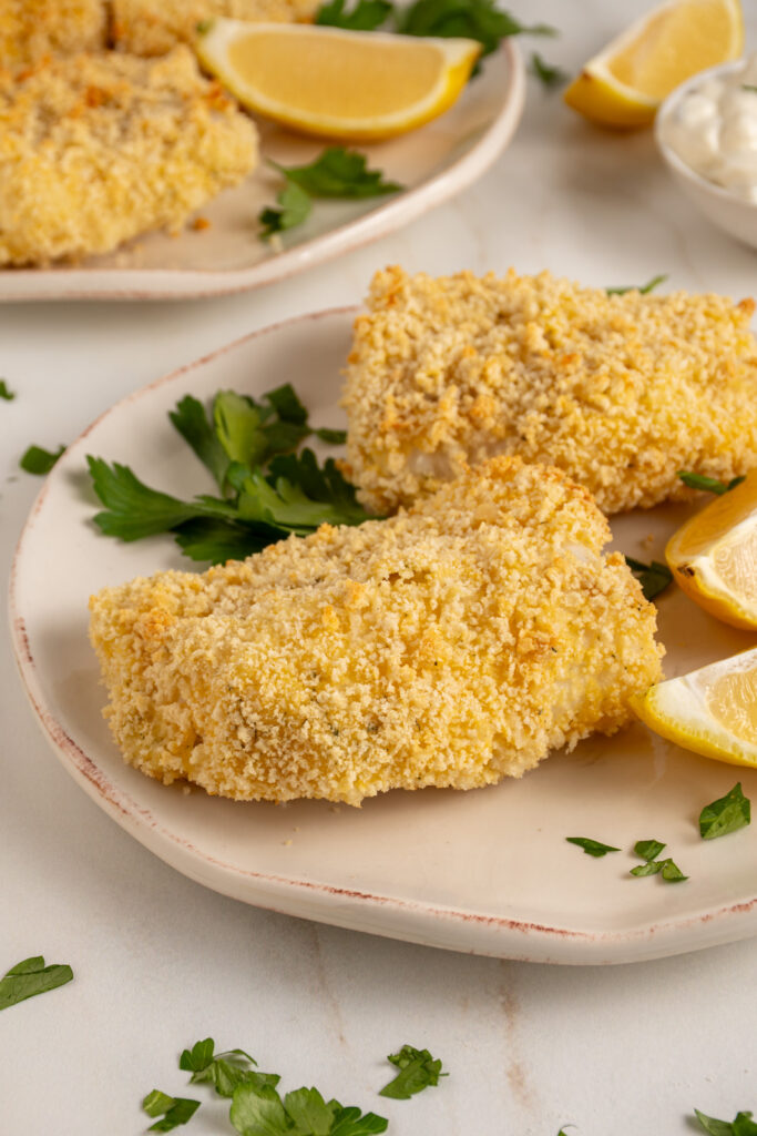 Plates with pieces of crispy baked breaded cod with lemons and fresh parsley.