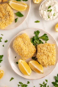 Plates with pieces of crispy baked breaded cod with lemons and fresh parsley.