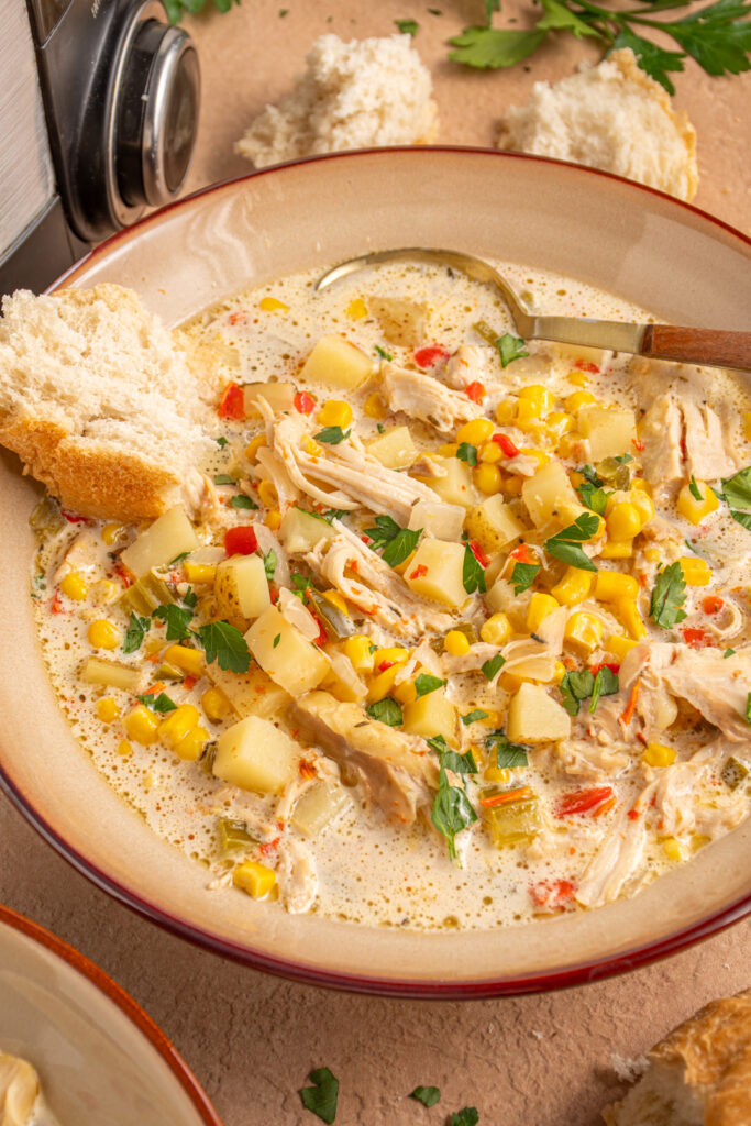 A brown bowl with chicken and corn chowder, a spoon, and a piece of crusty bread.