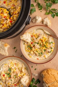 A brown bowl with chicken and corn chowder, a spoon, and a piece of crusty bread.