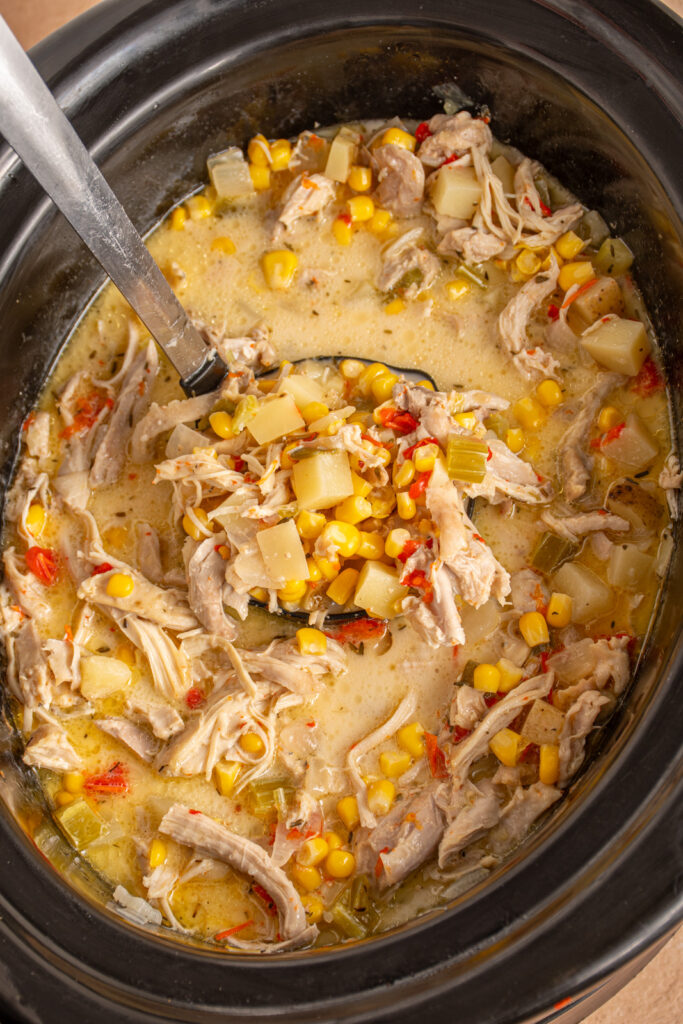 A black crockpot with chicken and corn chowder and a ladle.