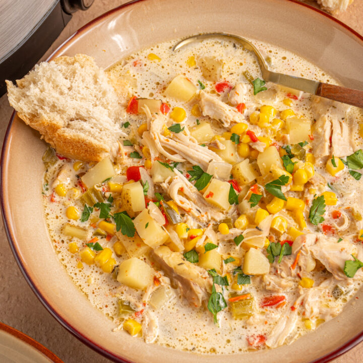 A brown bowl with chicken and corn chowder, a spoon, and a piece of crusty bread.
