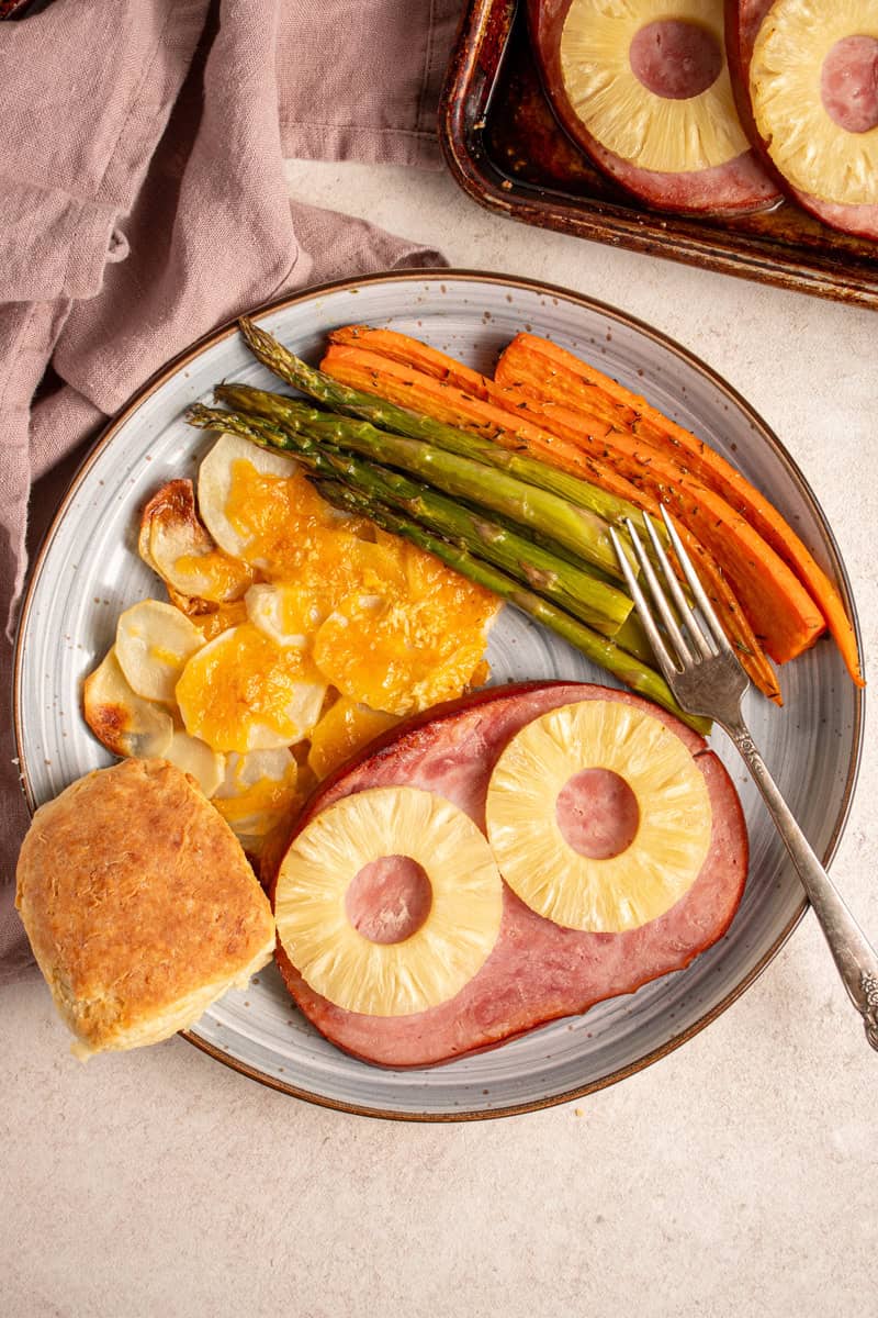 A plate with ham with pineapples, cheesy potatoes, asparagus, carrots, and biscuit.