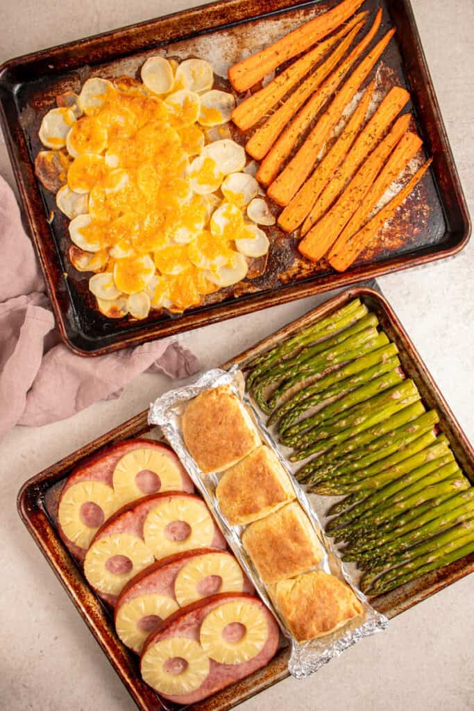 Two sheet pans filled with cooked ham, biscuits, asparagus, cheesy potatoes, and carrots.