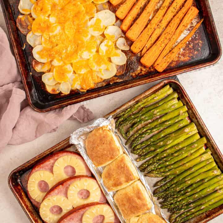 Two sheet pans filled with cooked ham, biscuits, asparagus, cheesy potatoes, and carrots.