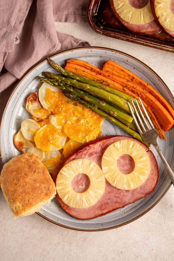 A plate with ham with pineapples, cheesy potatoes, asparagus, carrots, and biscuit.