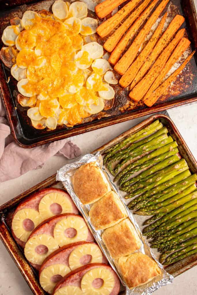 Two sheet pans with cooked ham and pineapple, biscuits, asparagus, cheesy potatoes, and carrots.