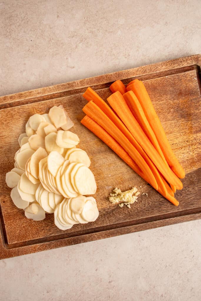 A cutting board with sliced potatoes, minced garlic, and halved carrots.