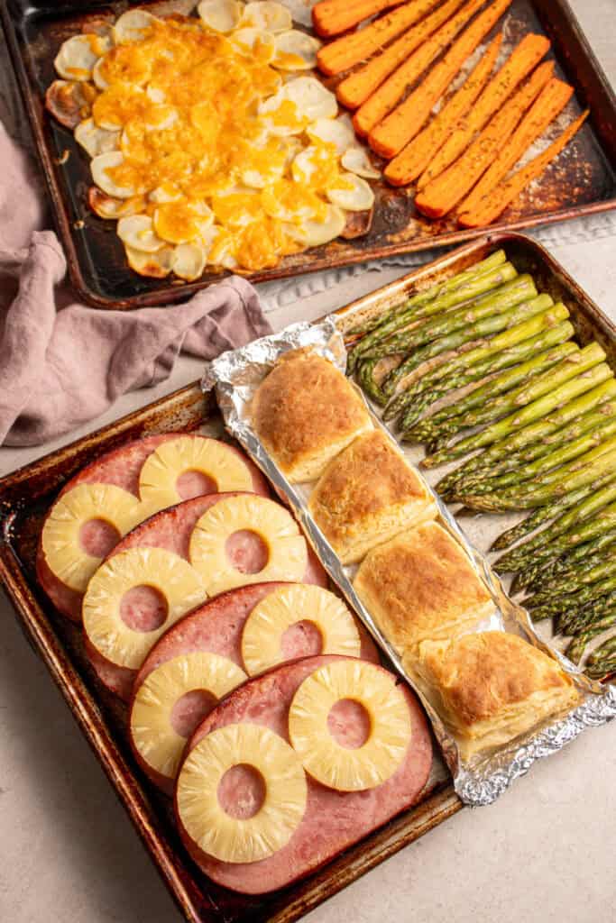 Two sheet pans filled with cooked ham, biscuits, asparagus, cheesy potatoes, and carrots.