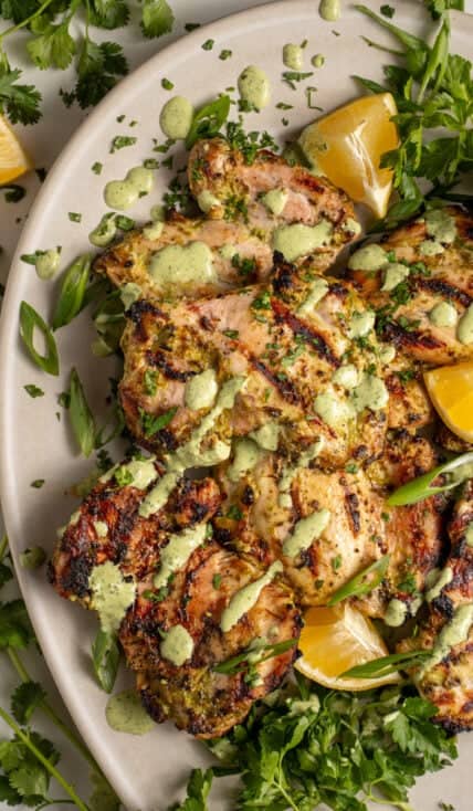 A platter of grilled green goddess chicken thighs with green goddess sauce and fresh herbs.