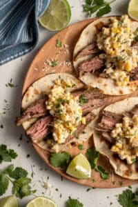 Steak and elote tacos laid out on a wood plate.