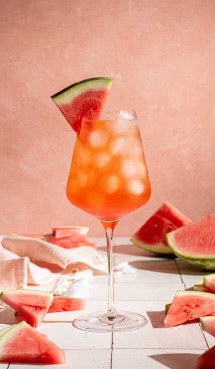 A wine glass filled with ice and Watermelon Aperol Spritz with a watermelon wedge.