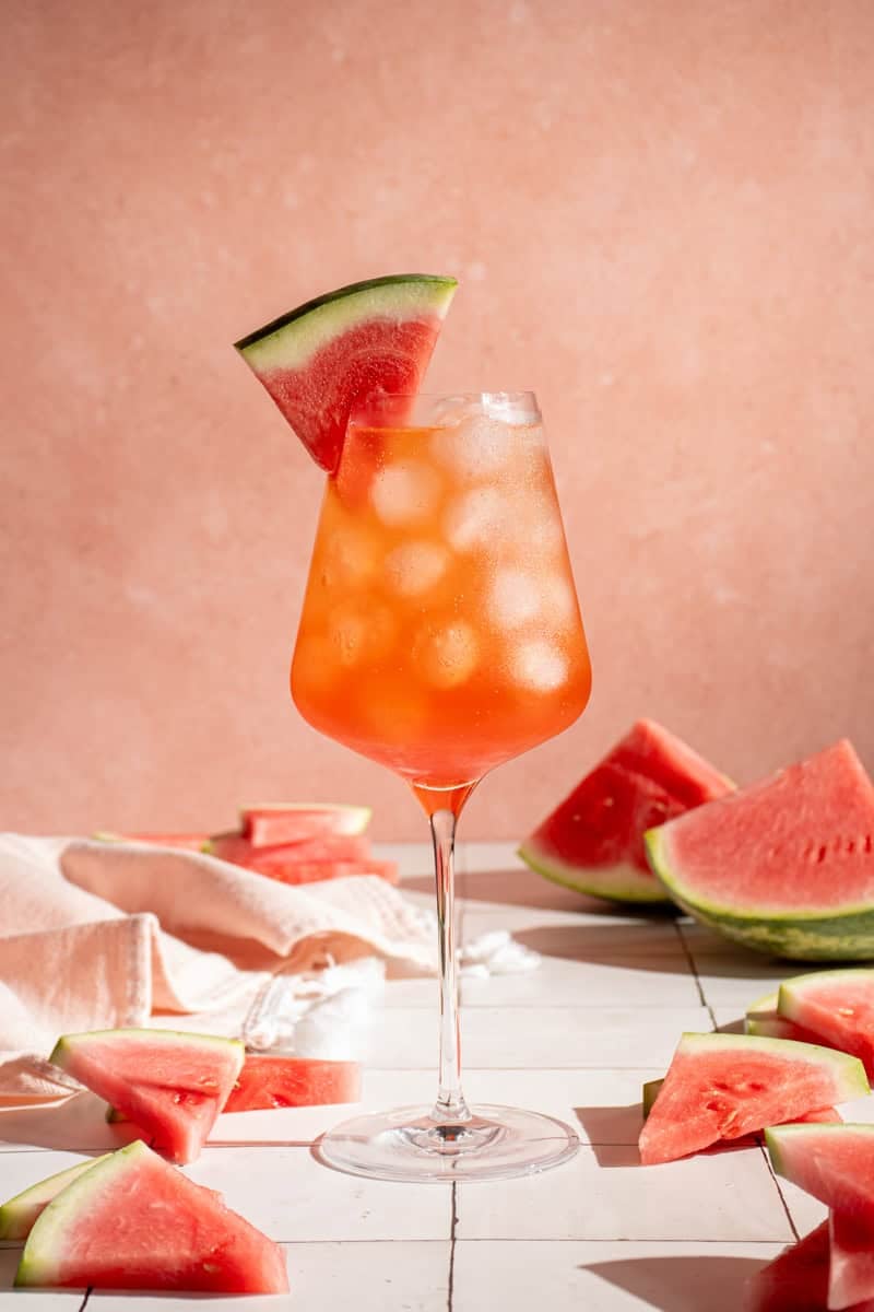 A wine glass filled with ice and Watermelon Aperol Spritz with a watermelon wedge.