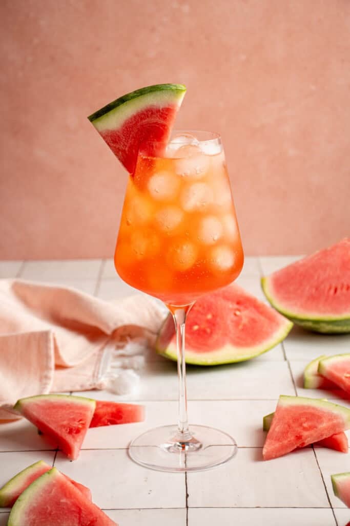 A wine glass filled with ice and Watermelon Aperol Spritz with a watermelon wedge.