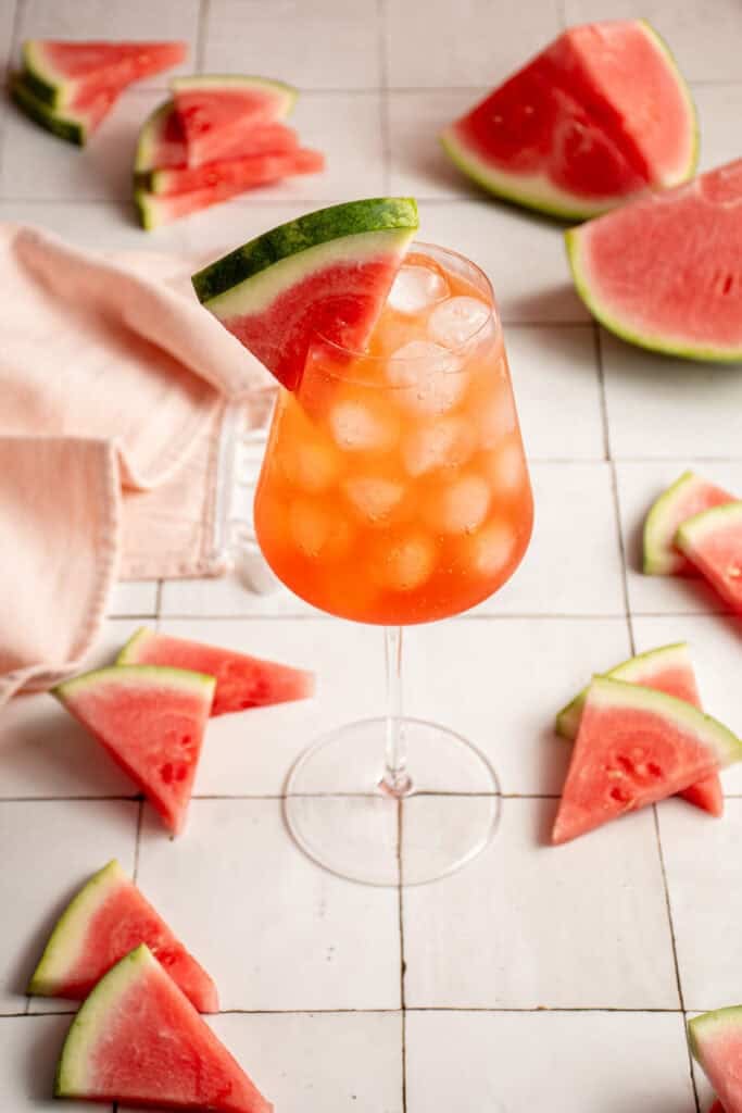 A wine glass filled with ice and Watermelon Aperol Spritz with a watermelon wedge.