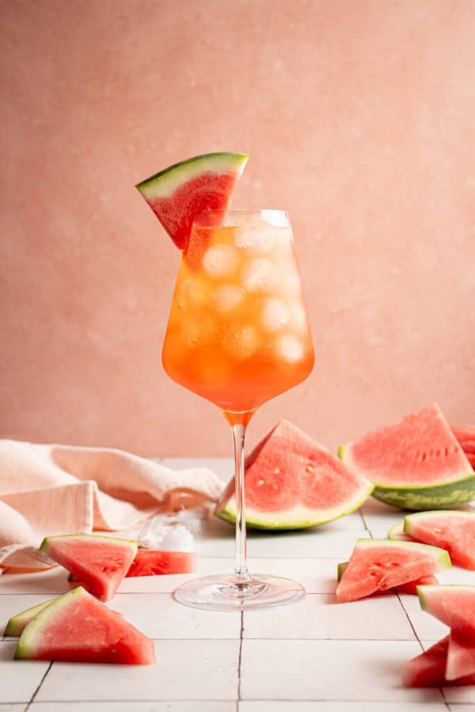 A wine glass filled with ice and Watermelon Aperol Spritz with a watermelon wedge.
